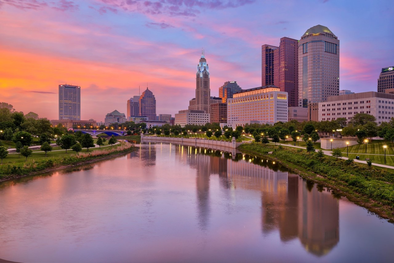 Image of downtown Columbus on the Scioto river at sunset. Giant river, tall buildings, and grassy parks with an orange, pink, blue sky illuminating the scene.